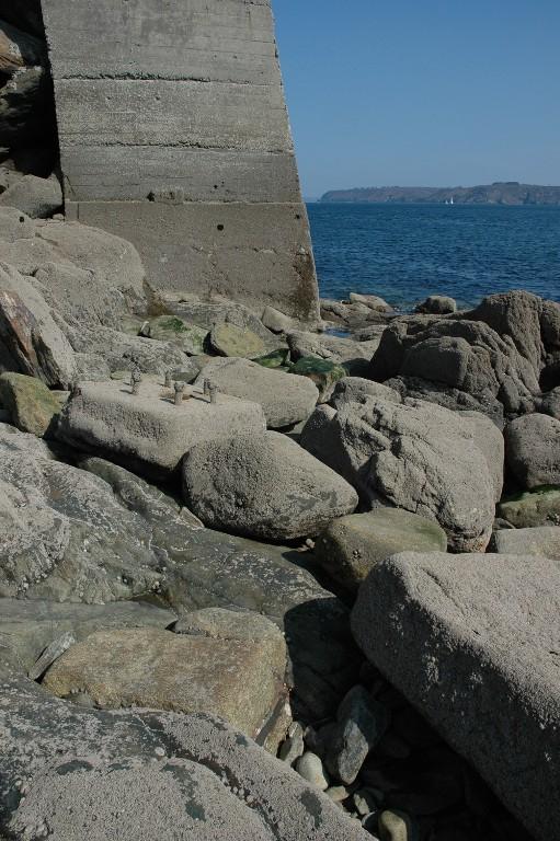Vue au premier plan des fondations de la batterie basse sur la grève à marée basse (état en 2005). Au centre, un dé d'ancrage en pierre de taille de granite à six tiges filetées pour une pièce d'artillerie. En arrière-plan, la partie consolidée en béton